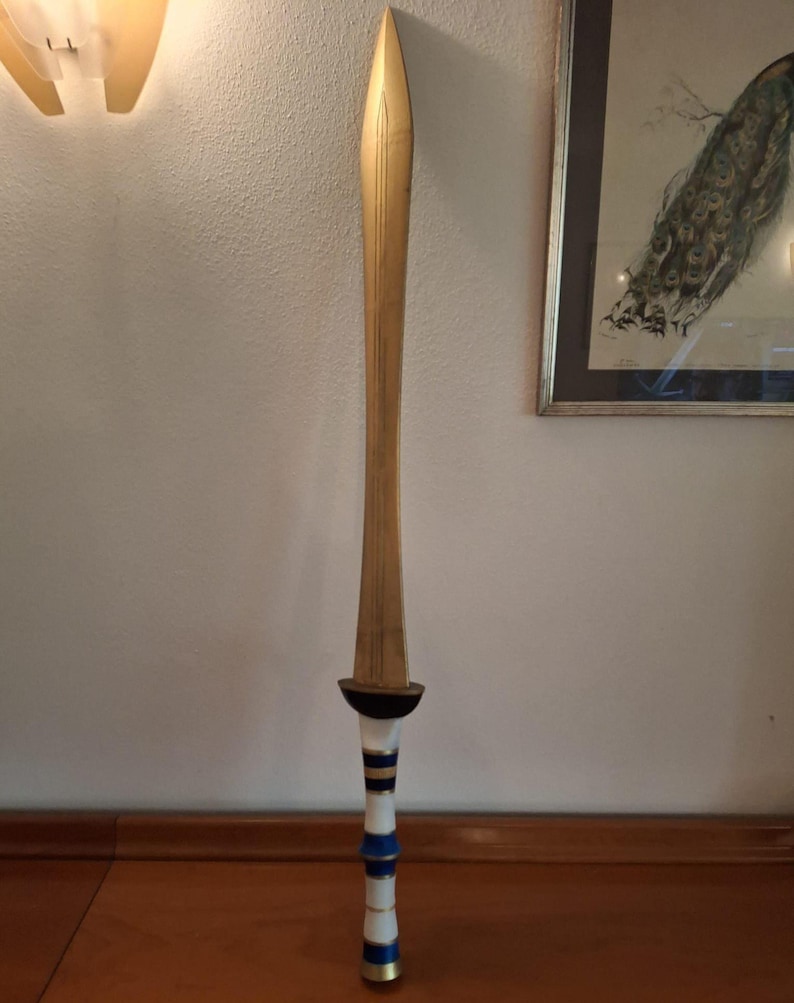 A tall fantasy sword with a golden blade and a hilt featuring white, blue, and gold bands standing vertically against a white wall.