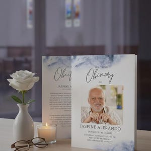 May include: A memorial service arrangement featuring a white rose in a small vase, a lit candle, and eyeglasses. Two obituary cards are displayed, one with the text "Obituary" and the other with a photo and the text "In Loving Obinary of JASPINE ALERANDO".