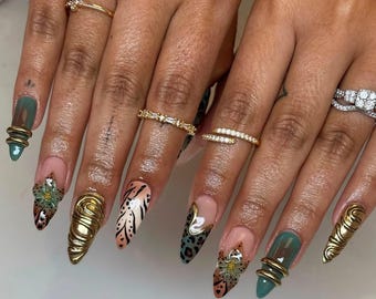 Puede incluir: Primer plano de uñas manicuradas con diversos diseños. Algunas uñas están pintadas de verde azulado, otras tienen una base nude con acentos florales y de estampado animal. Anillos de oro adornan los dedos, agregando un toque de elegancia.
