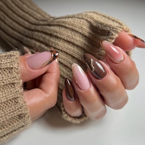May include: Long, oval-shaped nails in a palette of brown and pink, with gold detailing. Some nails are a single colour, others have gold outlines or abstract patterns. The manicure is well-executed, presenting a contemporary and sophisticated aesthetic.