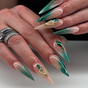 Op de afbeelding: Lange, puntige acrylnagels met een nude basis en een verloop van groen en wit. Sommige nagels hebben gouden en smaragdgroene versieringen. De nagels zijn een modieus accessoire.