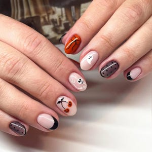 May include: A set of Halloween-themed nail art designs. Each nail features a different design, including a ghost, a bow, cherries, a skull, and polka dots. The nails are painted in various colors, including orange, black, and nude.