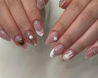 May include: A set of manicured nails with a light pink base and various designs. Some nails feature white tips with pearl accents, while others showcase 3D teddy bears, bows, and a white bunny. The nails have a glossy finish.