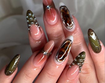 Puede incluir: Primer plano de una mano con uñas largas en forma de almendra. Las uñas presentan una variedad de diseños, incluyendo un degradado de rosa a marrón, un estampado de leopardo y verde oliva. Algunas uñas están adornadas con pedrería.