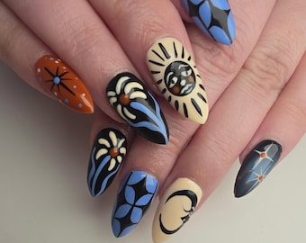 Op de afbeelding: Close-up van een hand met lange, amandelvormige nagels, elk beschilderd met unieke ontwerpen. De ontwerpen omvatten een zon, bloemen en geometrische patronen in kleuren als oranje, zwart, blauw en crème. De nagels zijn goed verzorgd en tonen nail art.
