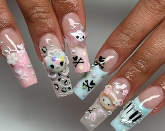 May include: A set of long, square-shaped acrylic nails with various designs. The nails feature 3D embellishments like a star, a cat, and a character, along with painted patterns, including skulls, flowers, and piano keys. Colors include pink, blue, white, and gray.