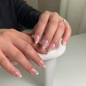 Könnte beinhalten: Nahaufnahme einer Hand mit French Manicure. Die Nägel sind mit einer hellrosa Basis und einer hellgelben Spitze lackiert. Ein silberner Ring mit einem großen, klaren Stein befindet sich am Ringfinger.