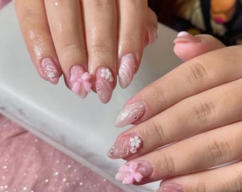 May include: Close-up of manicured nails with a pink and white color scheme. The nails feature 3D embellishments, including pink bows and white flowers. Some nails have a glittery finish, while others have a marble effect. The nails are oval-shaped.