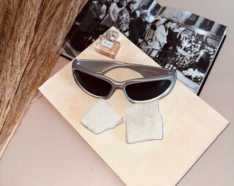 May include: Silver-framed sunglasses with dark lenses rest on a beige surface, next to a perfume bottle and a black and white photograph. The sunglasses have a sleek, modern design. The image is set against a soft pink background.