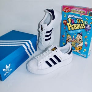 May include: White Adidas Superstar sneakers with black stripes and a white sole. A blue Adidas box and a box of Marshmallow Fruity Pebbles cereal are also in the image. The sneakers have the word "Superstar" on the side.