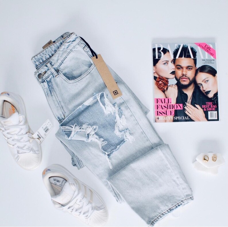 May include: Pale blue ripped jeans with a brown tag, white trainers, a fashion magazine, and gold earrings on a white surface. The magazine cover features models and the text "FALL FASHION ISSUE + BEAUTY SPECIAL".