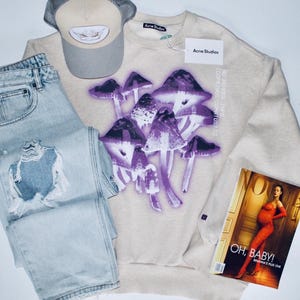 May include: A beige Acne Studios sweatshirt with a purple mushroom graphic, light wash denim jeans, and a beige trucker hat. A magazine with the text "OH, BABY!" is also visible.