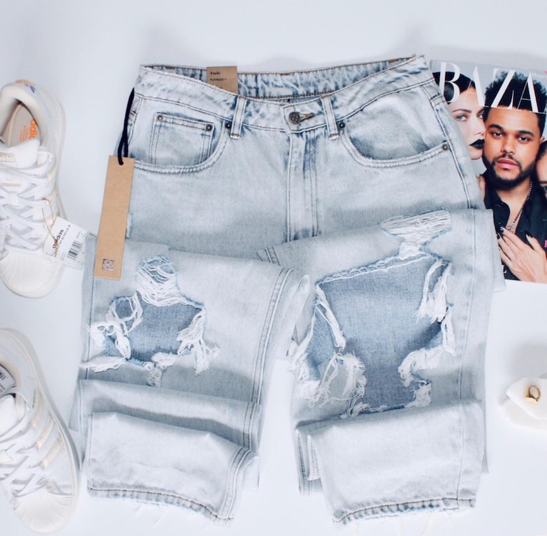 May include: Light blue distressed jeans with ripped details. The jeans have a classic five-pocket design and a button and zip closure. A brown tag hangs from the jeans. White trainers are visible in the background.