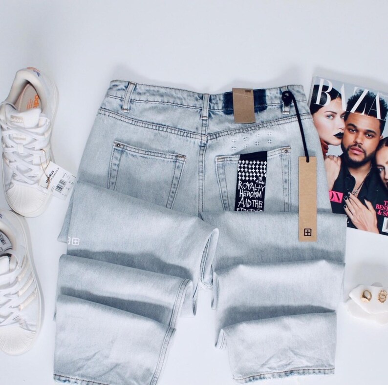 May include: Pale blue denim jeans with a faded wash, featuring a black and white tag that reads "ROYALTY HEROISM AND THE STREETS". The jeans are folded, with a brown tag attached. White trainers and a magazine are also visible.