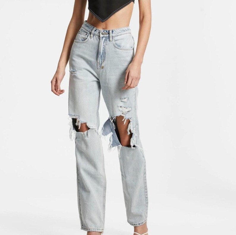 May include: Light wash denim jeans with distressed details, featuring large rips at the knees. The jeans have a classic five-pocket design and a straight leg fit. The light blue colour and ripped details give the jeans a casual, worn look.