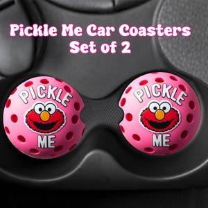 May include: Set of two pink car coasters with a cartoon character design. Each coaster has the words "PICKLE ME" and a red cartoon face. The coasters are designed to fit in a car's cup holders.