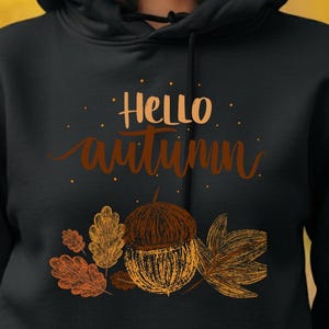Peut inclure: Un sweat à capuche noir avec l'inscription "Hello Autumn" en marron, ainsi qu'un motif de glands et de feuilles. Le design présente des couleurs d'automne sur un fond jaune flou.