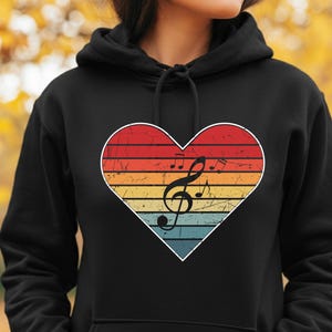 Op de afbeelding: Zwarte hoodie met een hartvormig ontwerp in een retro kleurenschema van rood, oranje, geel en blauw. Een witte omtrek omlijnt het hart, dat een zwarte G-sleutel en muzieknoten bevat.