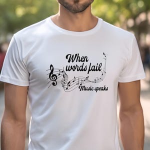 Könnte beinhalten: Weißes T-Shirt mit dem schwarzen Schriftzug "When words fail, Music speaks". Darüber befindet sich ein Notensystem mit Noten und einem Violinschlüssel. Das T-Shirt besteht aus weichem Material.