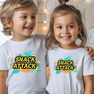 Op de afbeelding: Twee kinderen dragen witte t-shirts met de woorden "SNACK ATTACK" in geel, omlijnd met zwart, over een kleurrijk verfspatterontwerp. De kinderen lachen en lijken gelukkig.