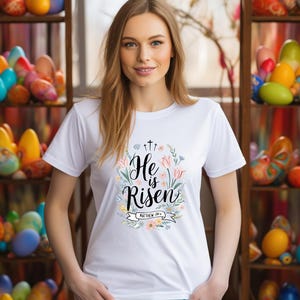 May include: White t-shirt with the words "He is Risen" in a floral wreath design. The design includes three crosses, flowers, and the scripture reference Matthew 28:6. The shirt is worn by a person with long blonde hair.