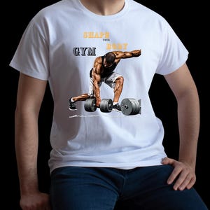 May include: White t-shirt featuring a weightlifter lifting dumbbells. The text "SHAPE YOUR BODY GYM" is printed in yellow and grey. The shirt is worn by a person in blue jeans.
