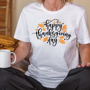 May include: A white t-shirt with the words "Happy Thanksgiving Day" in black script, surrounded by orange leaves and dots. The person is holding a white mug and wearing black trousers.