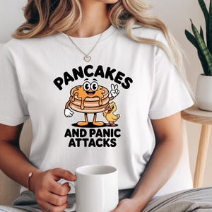 Może przedstawiać: Biała koszulka z postacią z kreskówki przedstawiającą naleśnik z symbolem pokoju, obok małej kaczki. Tekst na koszulce brzmi "PANCAKES AND PANIC ATTACKS". Projekt wykorzystuje brązowe, pomarańczowe i żółte kolory.