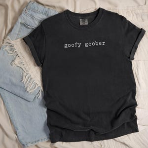 May include: Black t-shirt with the words "goofy goober" in white, paired with light blue jeans. The t-shirt has rolled-up sleeves and a vintage look. The jeans are frayed at the bottom.