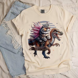May include: Cream-colored t-shirt featuring a whimsical illustration of a cat wearing sunglasses and a crown, riding a dinosaur. The cat has pink and purple feathered wings. The dinosaur is brown and blue. The shirt has a Comfort Colors tag.
