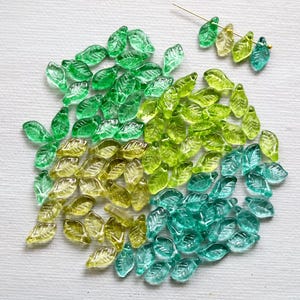 May include: A selection of glass leaf beads in shades of green, yellow, and teal. The beads are arranged on a white surface, with some strung on a gold wire. These beads are suitable for jewellery making and craft projects.