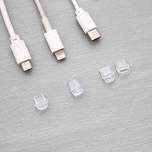 May include: Three white charging cables with different connector types: USB-C, Lightning, and Micro-USB. Also, there are four small, clear plastic cable protectors. The items are arranged on a light grey surface.