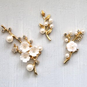 Könnte beinhalten: Drei goldfarbene Blumenbroschen mit weißen Blüten, Kunstperlen und klaren Kristallen. Die Broschen sind in Form von Zweigen mit Blättern und Blüten gestaltet. Der Hintergrund ist hellgrau.