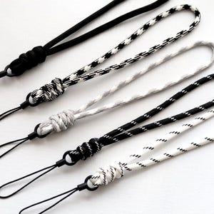 Könnte beinhalten: Eine Sammlung von fünf Handgelenksschlaufen in verschiedenen Mustern und Farben. Die Schlaufen verfügen über schwarze, weiße und graue seilartige Kordeln mit einer Schlaufe zur Befestigung an einem Telefon oder einem anderen Gerät. Jede Schlaufe hat ein Knotendetail.