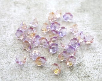 May include: A collection of small, translucent flower-shaped beads. The beads have a gradient of colors, including purple, peach, and yellow. They are scattered on a light gray surface, creating a delicate and whimsical aesthetic.