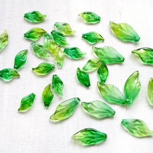 May include: A collection of translucent green glass leaf beads in various sizes and shades, scattered on a white surface. The leaves range from light yellow-green to deep emerald, with delicate veining details.