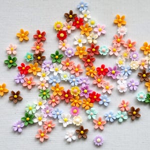 May include: A collection of small, colorful flower embellishments in various shades of orange, red, yellow, pink, purple, green, and brown. These miniature flowers have a detailed design and are scattered across a white surface, perfect for crafts.
