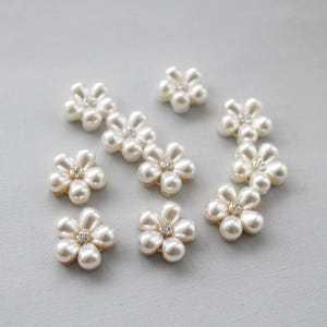 May include: A collection of off-white, pearl-like flower embellishments with a small, sparkling rhinestone center. The petals are rounded and arranged in a floral pattern. These decorative elements are ideal for craft projects.