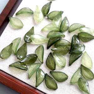 May include: A collection of small, translucent leaf-shaped beads in various shades of olive and sage green. Each bead features a smooth, curved surface with a subtle vein pattern and a small hole at the narrow end for stringing or jewelry making.