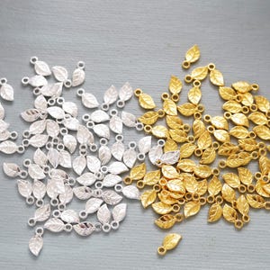 Könnte beinhalten: Eine Sammlung kleiner, blattförmiger Anhänger in Silber- und Goldtönen. Jeder Anhänger hat eine Schlaufe zum einfachen Anbringen an Schmuck oder anderen Bastelarbeiten. Die Anhänger sind auf einer grauen Oberfläche verstreut.
