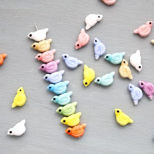 Peut inclure: Une collection de petites perles en forme d'oiseaux colorés, dans des tons d'orange, de jaune, de rose, de violet, de bleu, de vert et de blanc. Chaque perle a un petit point noir pour l'œil. Les perles sont dispersées sur une surface gris clair.