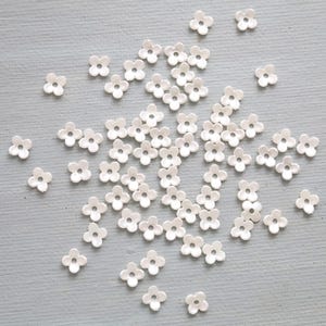 May include: A collection of small, white, flower-shaped embellishments scattered on a light gray surface. Each flower has five petals and a small hole in the center. The embellishments are likely used for crafts or decorations.