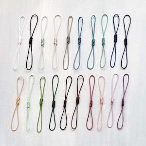 Peut inclure: Une collection de boucles de cordon nouées colorées dans diverses teintes, dont blanc, noir, beige, vert, bleu et violet. Les boucles sont disposées en rangées sur une surface blanche, présentant une gamme de couleurs et de textures.