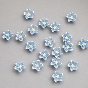 May include: A scattering of small, light blue flower-shaped embellishments with white centres. These decorative floral elements are displayed on a grey surface, highlighting their delicate details and design.