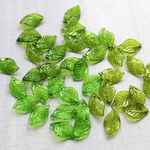 May include: A scattering of translucent green leaf-shaped beads. Each bead has a small hole for stringing. The beads are displayed on a white surface, highlighting their delicate details and varying shades of green.
