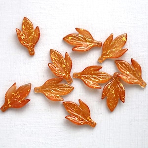 May include: A collection of ten small, translucent orange leaf-shaped charms. Each charm features a smooth, glossy finish with embedded gold-colored glitter flecks throughout the surface. Each leaf has a small hole at the base for attachment.