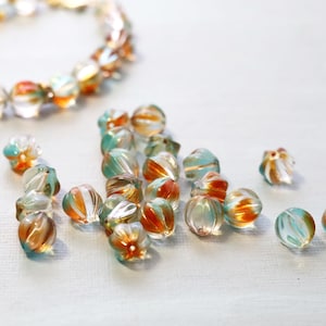 May include: A collection of round, translucent glass beads featuring a swirling pattern of orange, teal, and clear glass. The beads are arranged in a loose pile, with a circular bracelet made of the same beads positioned in the upper left corner.