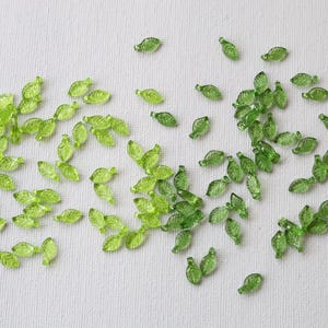 May include: Numerous small, translucent green glass leaf beads scattered on a white surface. Each leaf is detailed with veining and has a rounded shape. The beads create a delicate, natural aesthetic, suitable for crafting or jewellery making.