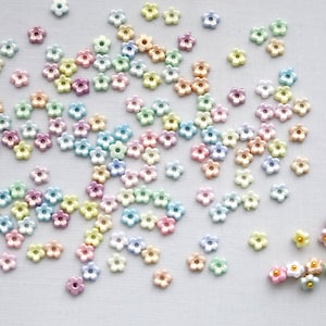 May include: A scattering of small, pastel-colored flower-shaped beads. The beads are in shades of pink, yellow, blue, green, and peach. Each bead has five petals and a small hole in the center. They are scattered on a white surface.