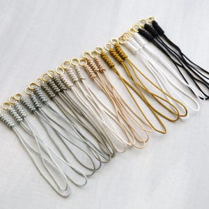 May include: A collection of handmade wrist straps in various colors, including gray, beige, gold, white, and black. Each strap features a knotted design and a gold-colored metal ring. The straps are arranged on a light-colored surface.